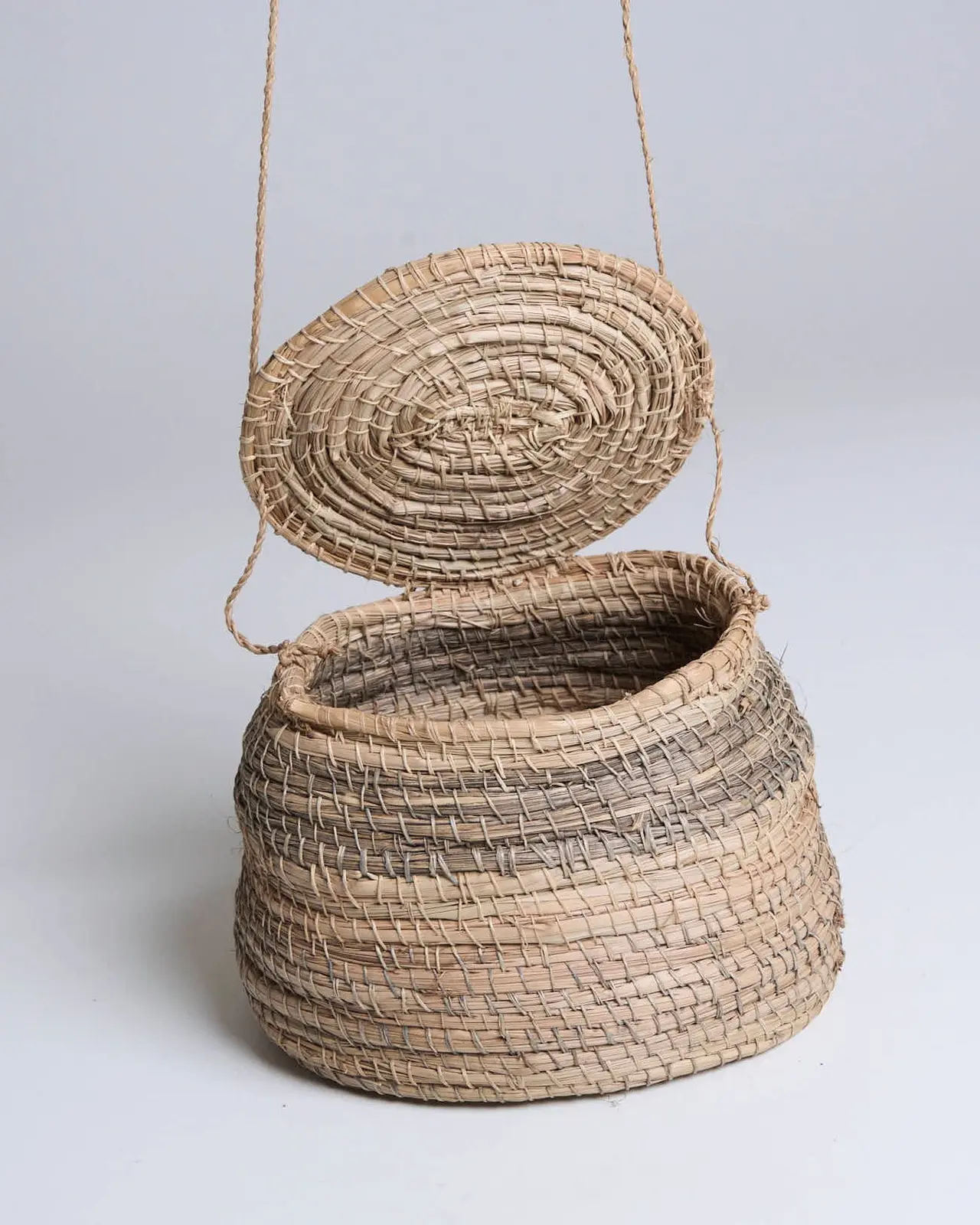 Bolsa arredondada de fibra bege, aberta, com tampa circular e alça fina.