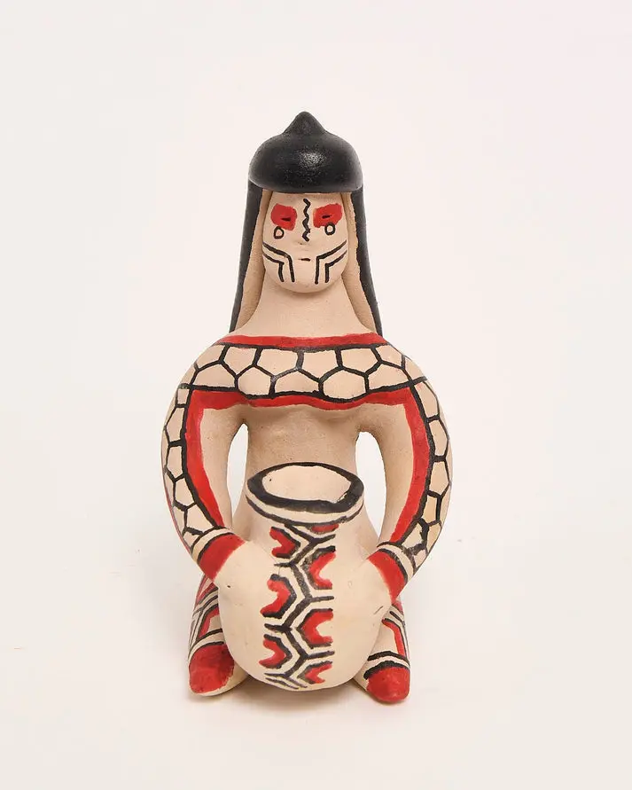 Boneca de cerâmica do povo Karajá, com cabelo preto longo, grafismos vermelhos e pretos e um pequeno pote nas mãos.